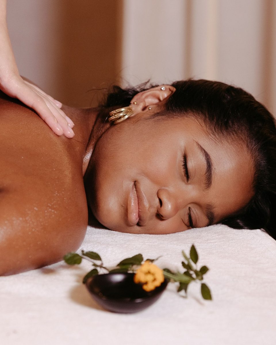 A woman smiles with her eyes closed as a massage therapist&rsquo;s hands work on her shoulder